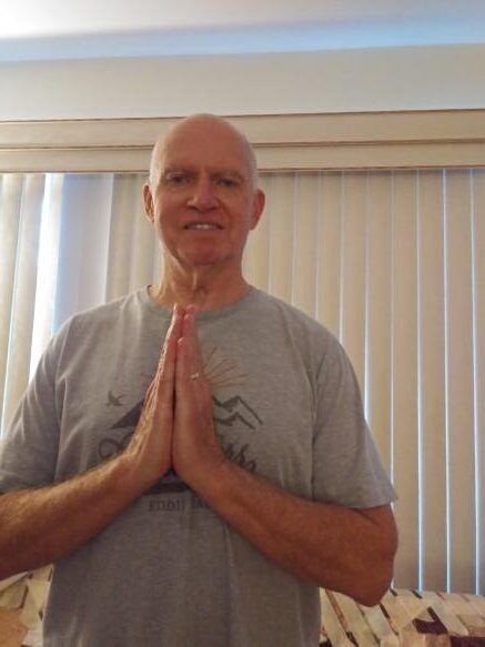 Charles Baber wearing a grey shirt and smiling with hands in a prayer position